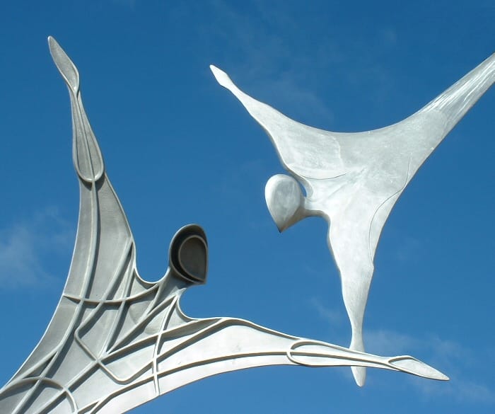 Image of a metal sculpture of two abstract figures seemingly reaching out to support each other. 